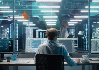 Photo from behind a man working at dual monitor workstation with coding on screen. In the background a woman walks in data center area, behind glass. Textured pattern overlay can be seen with orange diamond shape near upper left corner.