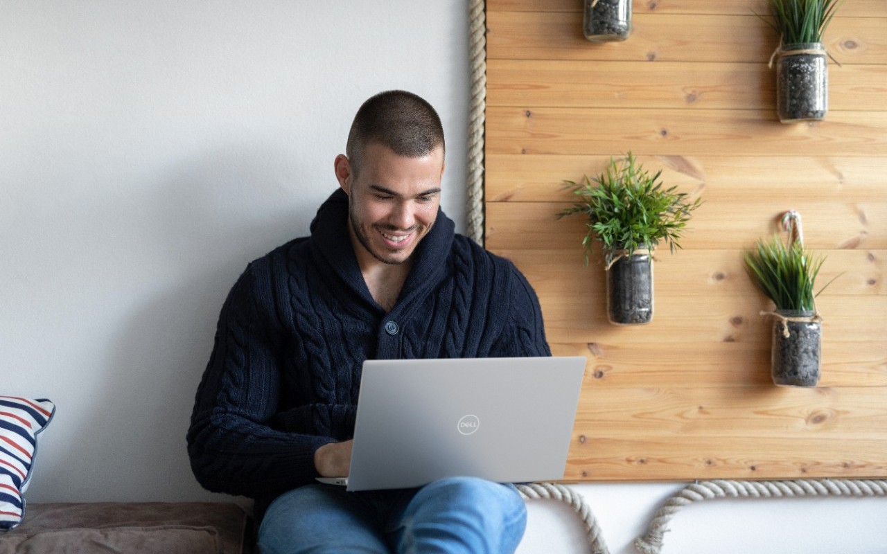 Man in blue jeans and hoodie using Dell Inspiron laptop on a bench with plants in the background.