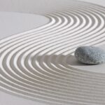 Japanese Zen garden close up of white sand raked into a curve pattern around a small gray stone.