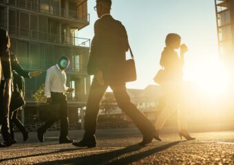 Business people walking on a busy street in the morning with a sun flare in the background.