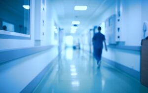 Health care professional walking down hospital corridor, away from camera, in defocused image, tinted in blue.