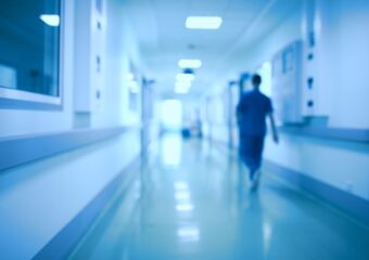 Health care professional walking down hospital corridor, away from camera, in defocused image, tinted in blue.