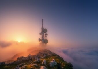 Telecommunications tower on a mountain top, above cloud layer with sun in background.