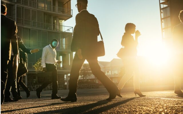 Business people walking on a busy street in the morning with a sun flare in the background.