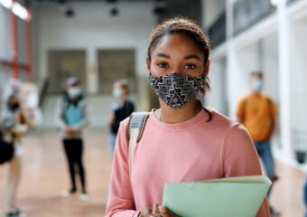 Young African-American female student at school, wearing facial covering as other students in the background also wear facial coverings.