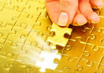 Woman holding gold color puzzle piece over spot in puzzle where it fits.