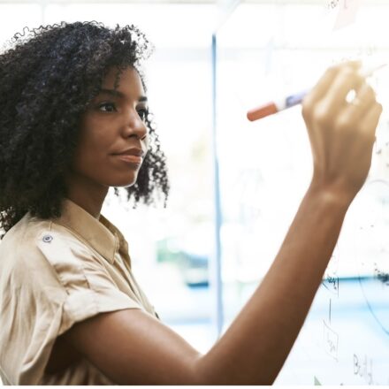 African-American woman writing out team roadmap plans on glass window with marker.