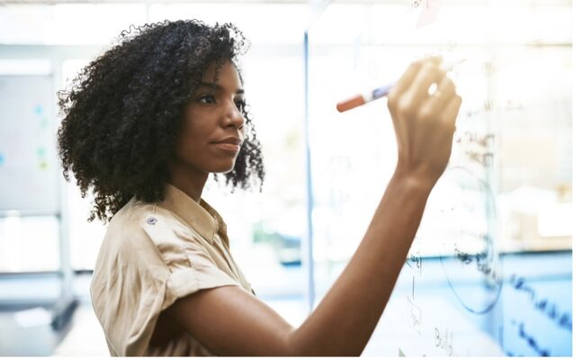 African-American woman writing out team roadmap plans on glass window with marker.