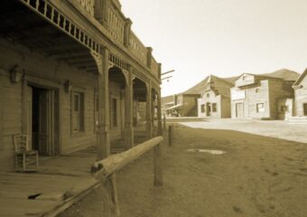 Image of Old West American town, dusty street, hitching post and wooden buildings, in sepia tone.
