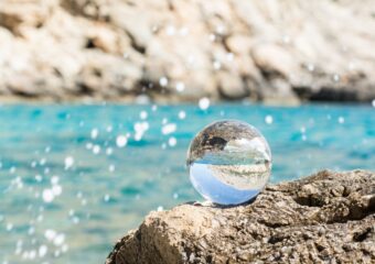 A clear glass ball showing sea and rocky shore through it as it sits on a rock outcropping along the Mediterranean Sea.