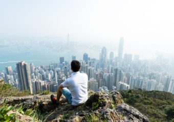 Man sitting on side of slope overlooking cityscape below.