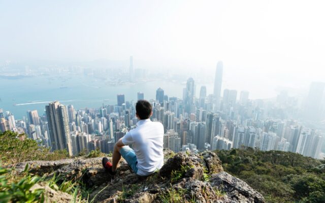 Man sitting on side of slope overlooking cityscape below.