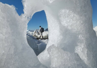 Person in a cold, snowy and icy outdoors environment carries a Dell Rugged Latitude notebook. Image of person taken through a hole in snow and ice formation with a blue sky in the background.