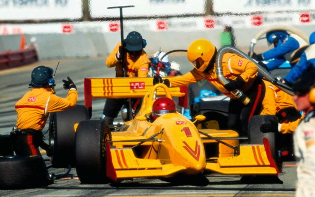 F1 race car team in yellow outfits work on the yellow race car with red stylized design during a pit stop.