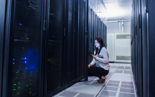 A female data center technician is kneeling next to a server rack, checking on data results on her laptop.