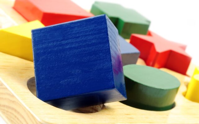 Blue, green, red, and yellow blocks of different shapes on a wodden toy surface with corresponding shaped holes.