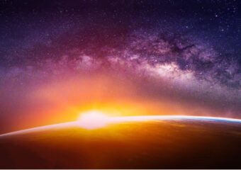 Digitally rendered image of sun emerging over Earth's horizon, light breaking and Milky Way galaxy in background. Image created digitally based on elements furnished by NASA.