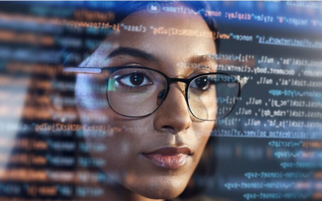 African-American woman wearing glasses reviews software coding. Image POV is from behind the coding looking out from inside the monitor.