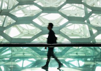 Defocused image of a man walking in a modern design corridor, with a hexagonal design wall or building section appears in the background.