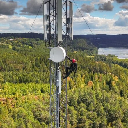 Field technician scales telecommunications tower with a forest and body of water in the background.