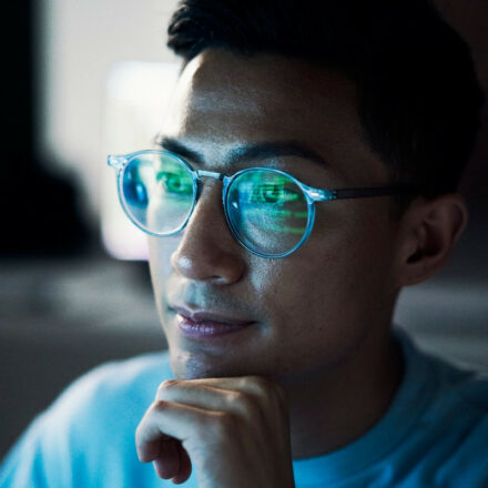 Asian man with glasses works on coding, with reflection of monitor appearing on his glasses.