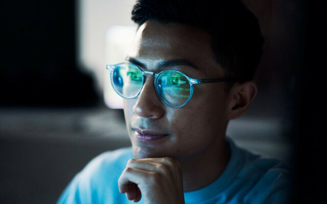 Asian man with glasses works on coding, with reflection of monitor appearing on his glasses.