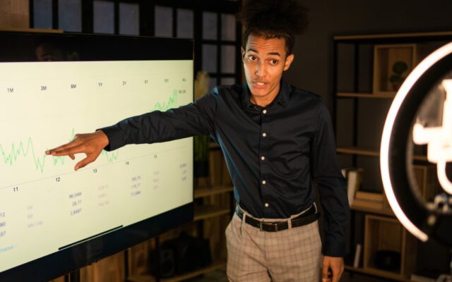 Young black man in business casual attire speaks to data on large screen for presentation.